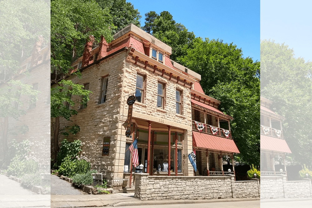 Historic stone building with red roof and American flag, set against lush greenery. The facade displays "Museum" signs and patriotic buntings. Sunny day.