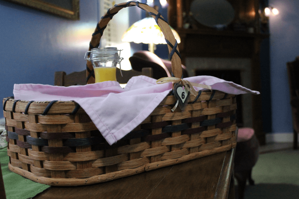 A woven basket with a pink napkin and a glass jar of juice sits on a wooden table in a cozy room.