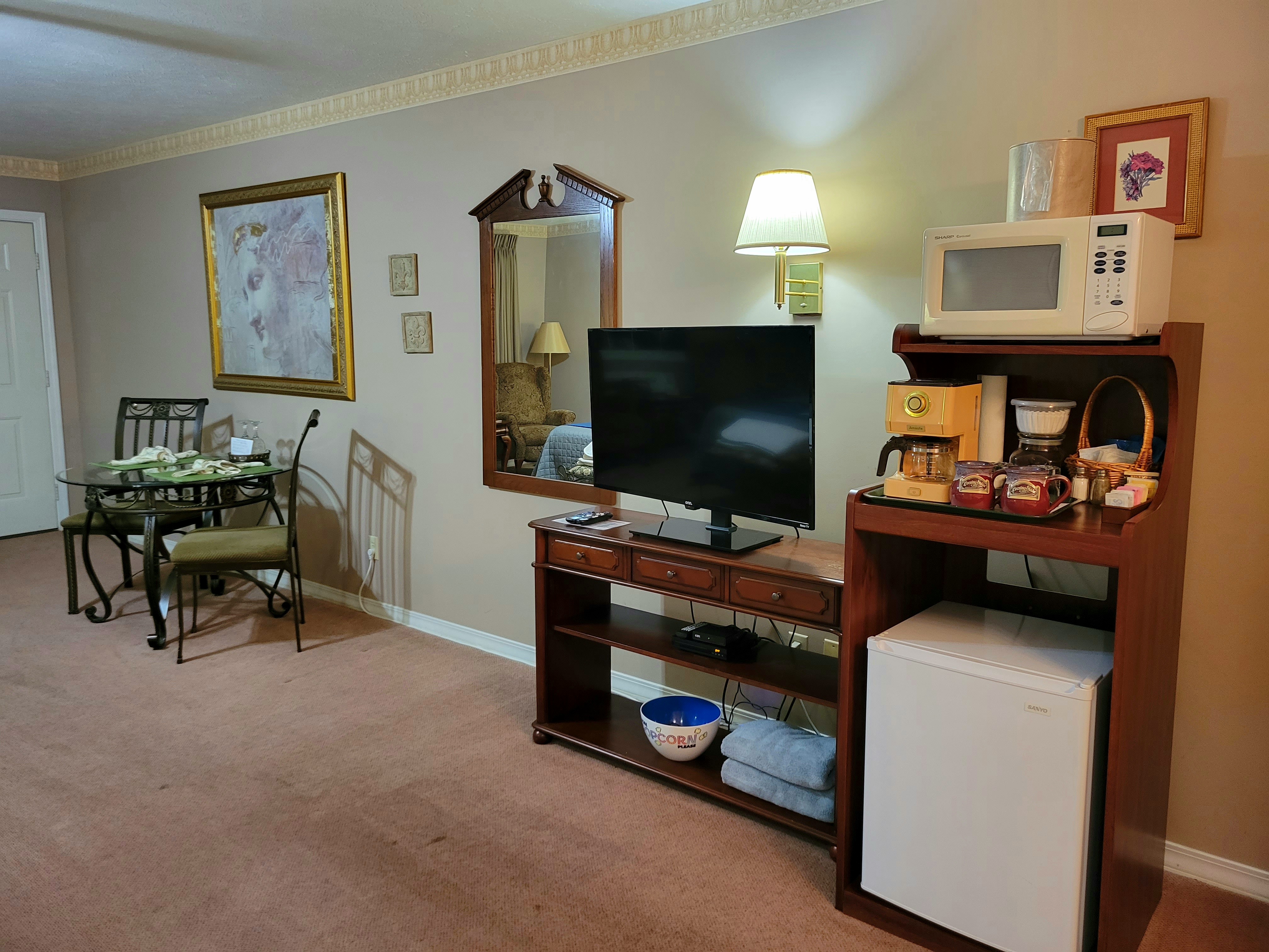 A suite showing a entertainment and refreshment center. A flat-screen TV on a wooden stand sits next to a kitchenette station equipped with a microwave, a vintage-style coffee maker, and a mini-fridge. In the background, a glass-top dining table and chairs are positioned under a large gold-framed artwork.