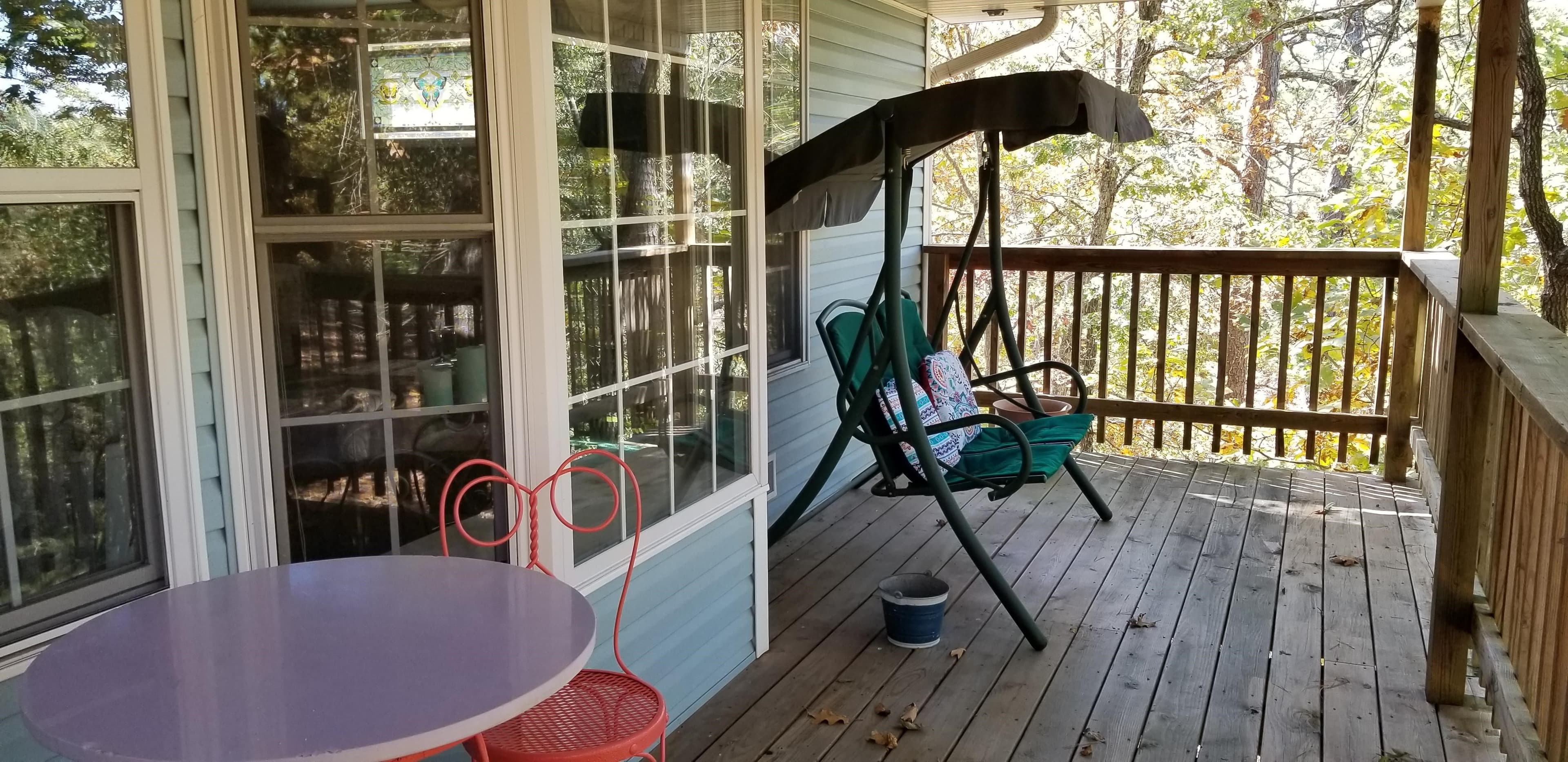 Exterior deck out front of the Dahlia cottage. Protruding out is the exterior portion of the bay window. In the left of the foreground is a white topped 2 person cafe table with salmon colored iron chairs. There is a forest green, 2 person swing on the deck in the background. The deck is a regular, treated mid brown wood color. There are woods off in the distance.