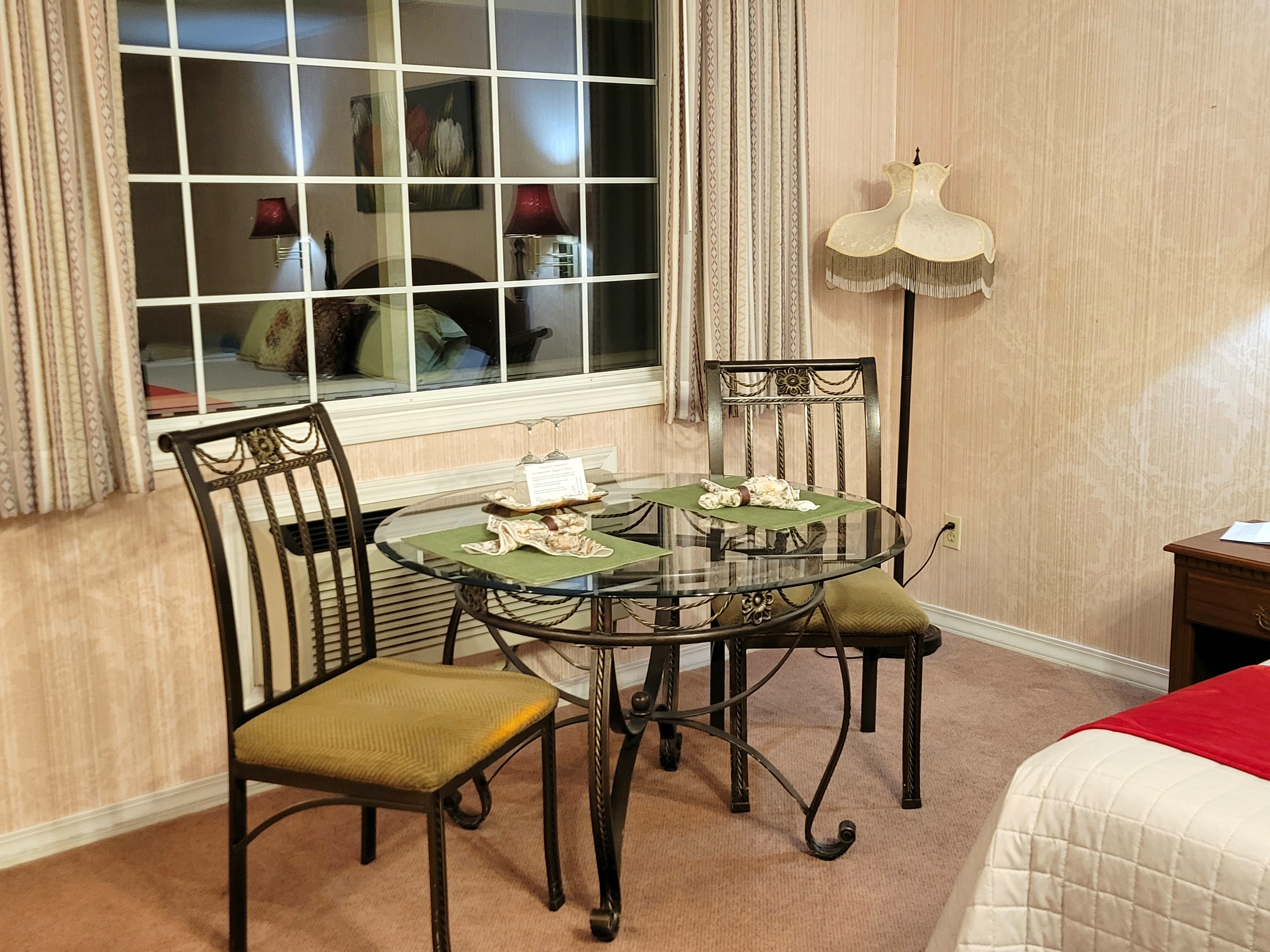 Glass table with 2 wine glasses, cloth napkins, and place settings,. 2 metal chairs with padded seats in front of a spacious opened window. Window view looks out on a paved parking lot and wooded Ozark treeline.