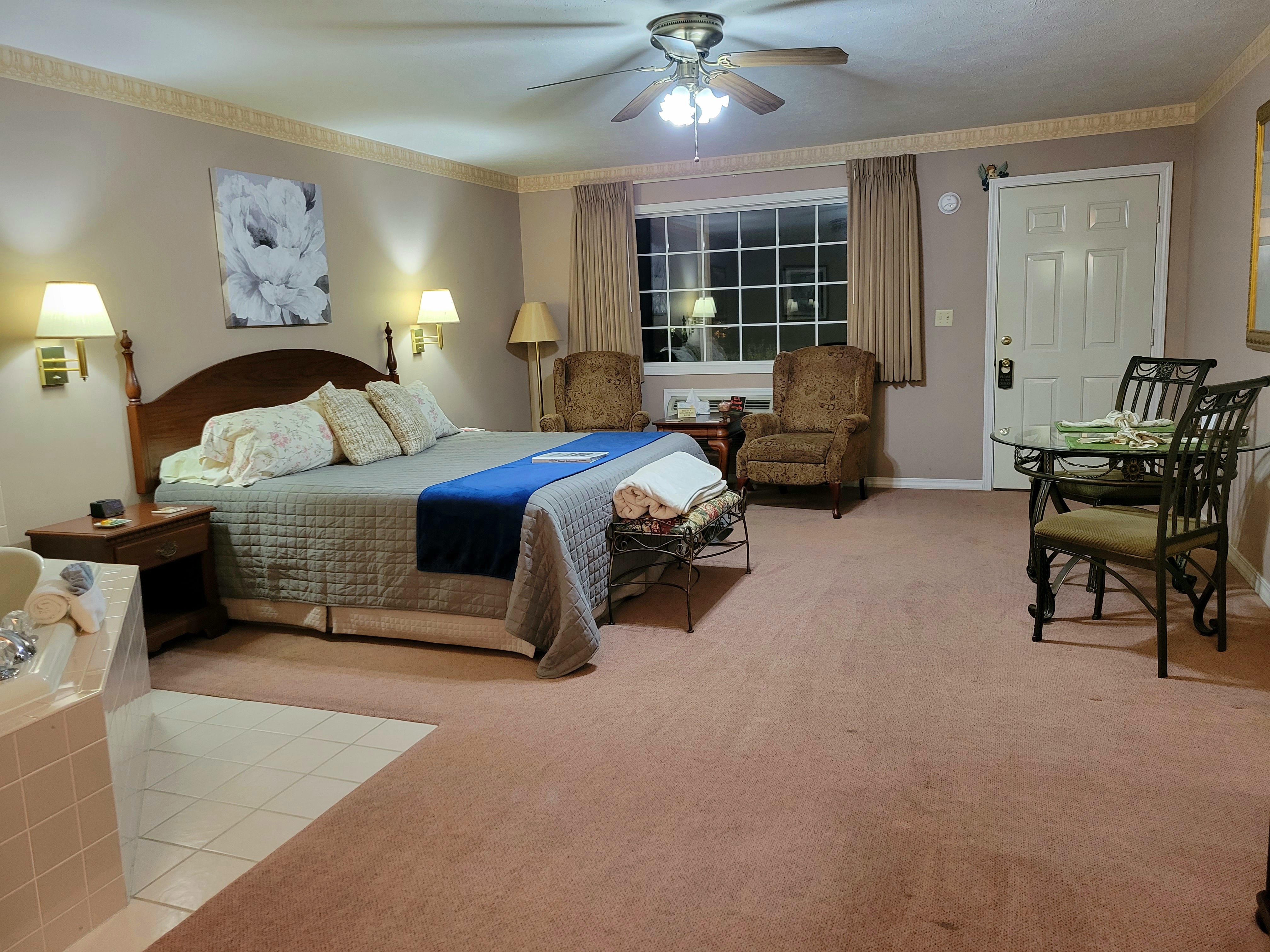 An interior view of a spacious suite featuring a king-size bed with a blue runner, two cozy armchairs by a large window, a dining table with chairs, and a ceiling fan. A large jetted tub is partially visible in the foreground.