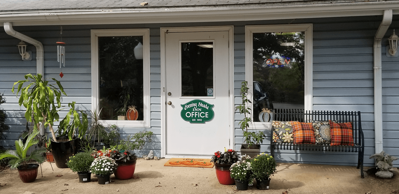 The entrance to an inn office with potted plants, a white door, a green office sign, bench with cushions, and an "open" sign in the window.
