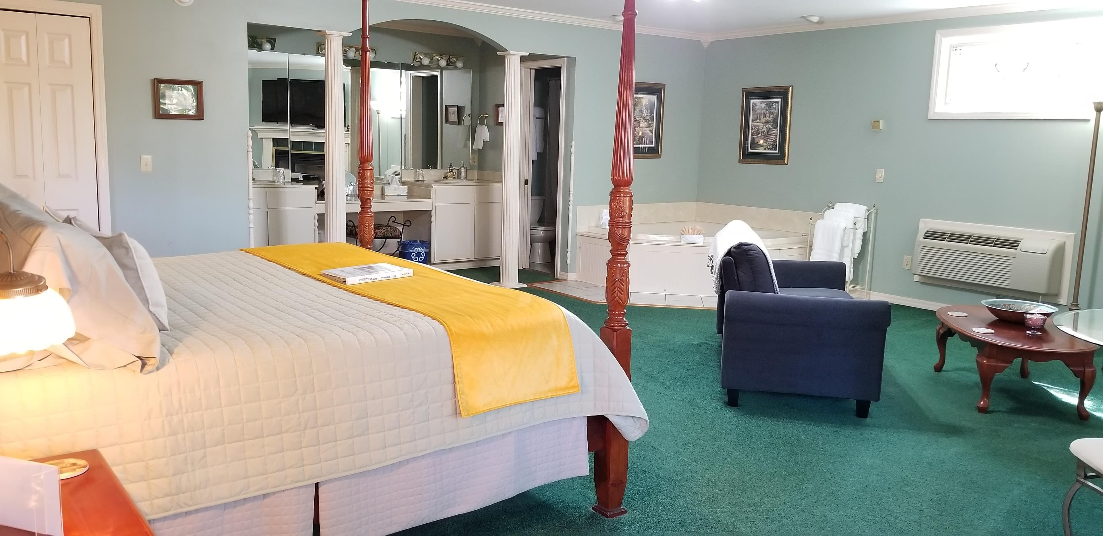Foot of 4 poster, wooden bed. Room has green carpet. Jetted tub and bathroom and vanity in the left side background. The walls are a light blue. There is a dark blue, velvet loveseat with a coffee table in front of it. The vanity area is recessed and has pillars in front in the arched doorway. The pillars and room trim are all in white.