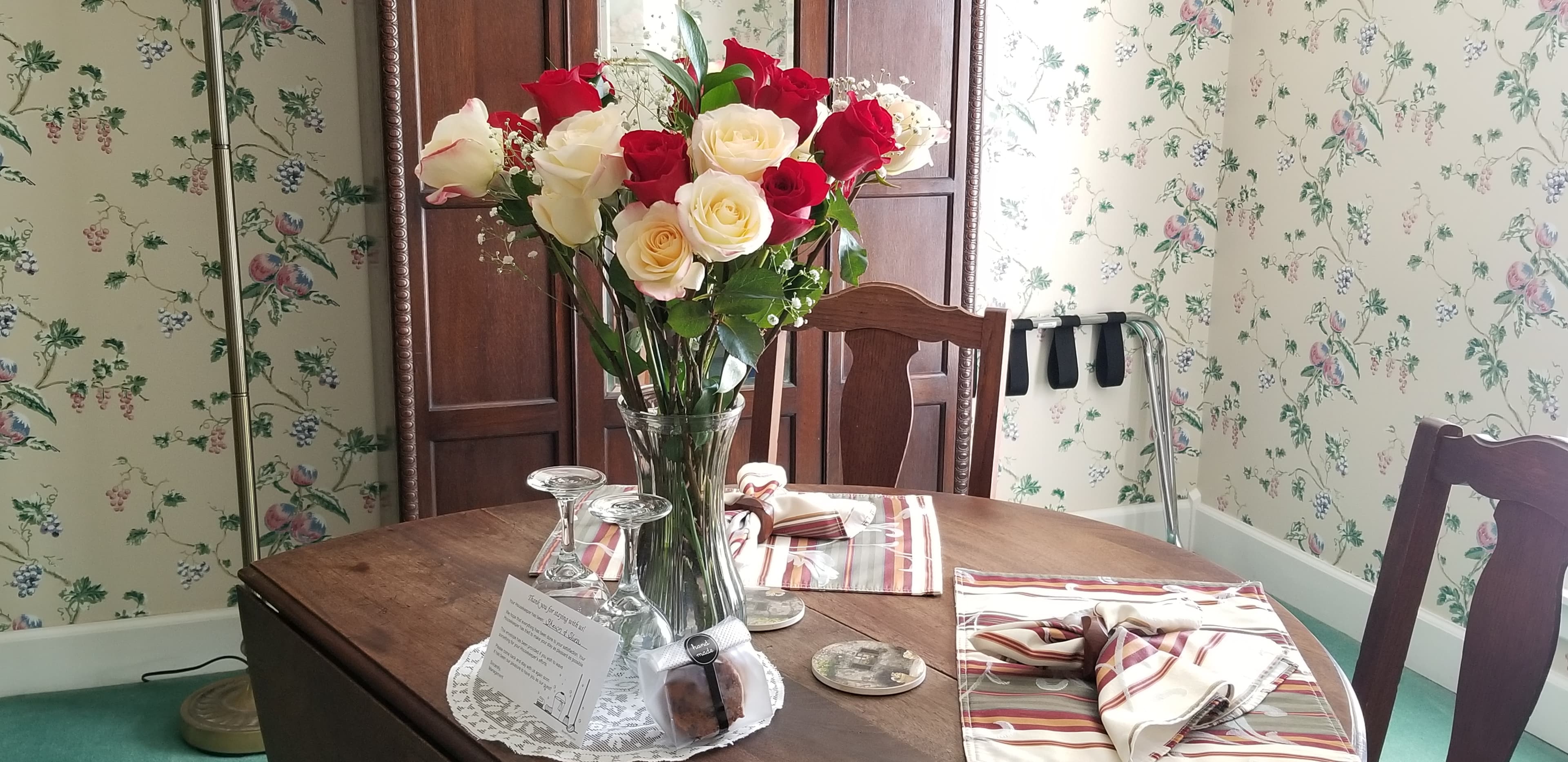 A dining table adorned with a large vase of red and white roses. The table is set with wine glasses and patterned napkins, positioned in front of a wooden armoire and floral-patterned wallpaper.
