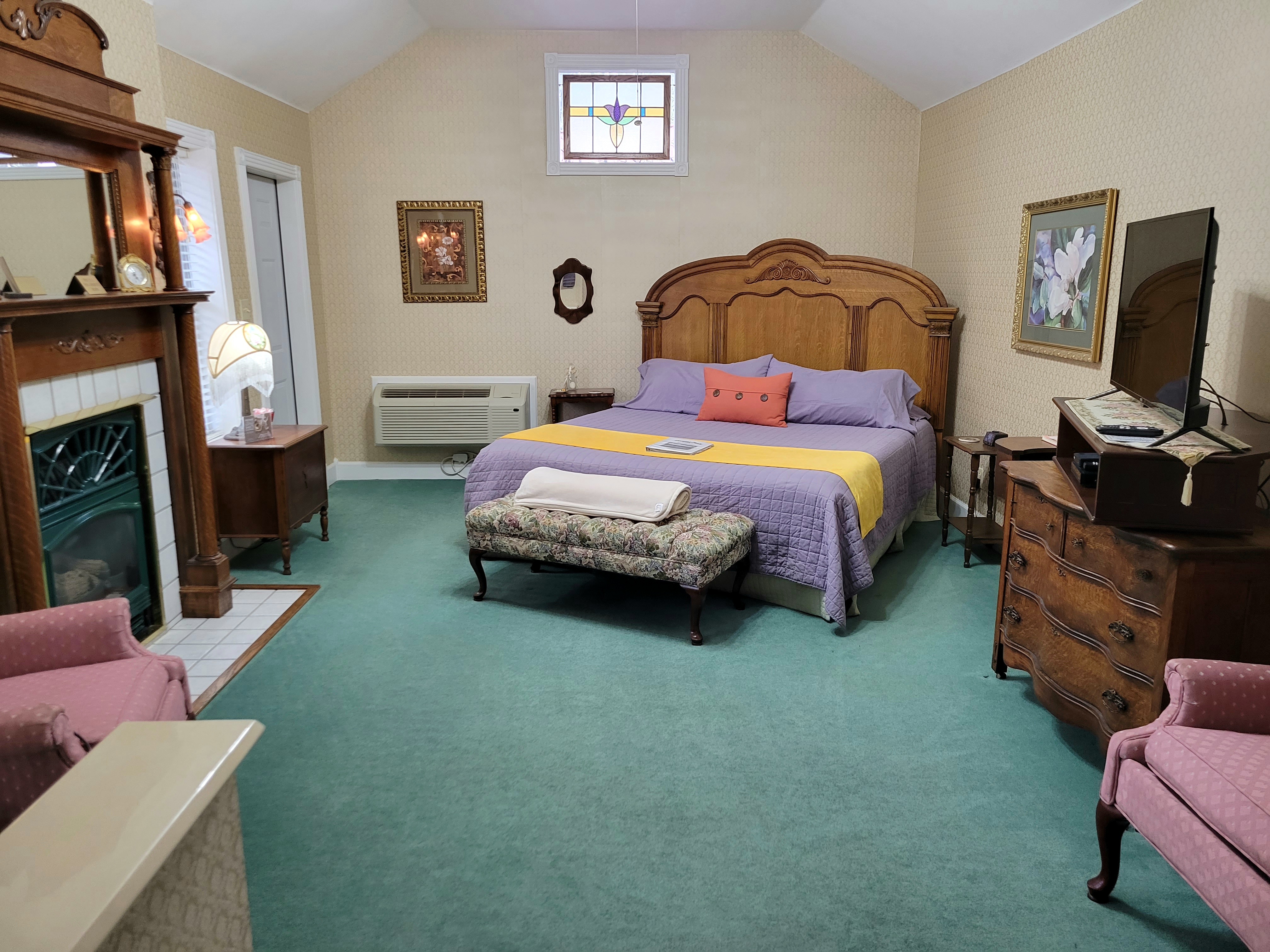 An interior view of a spacious bedroom featuring a large, ornate wooden king-size bed with purple linens and a yellow runner. The room is decorated in a vintage style with teal carpeting, two pink armchairs, a classic fireplace with a detailed wooden mantel, and a stained-glass window accent above the bed.