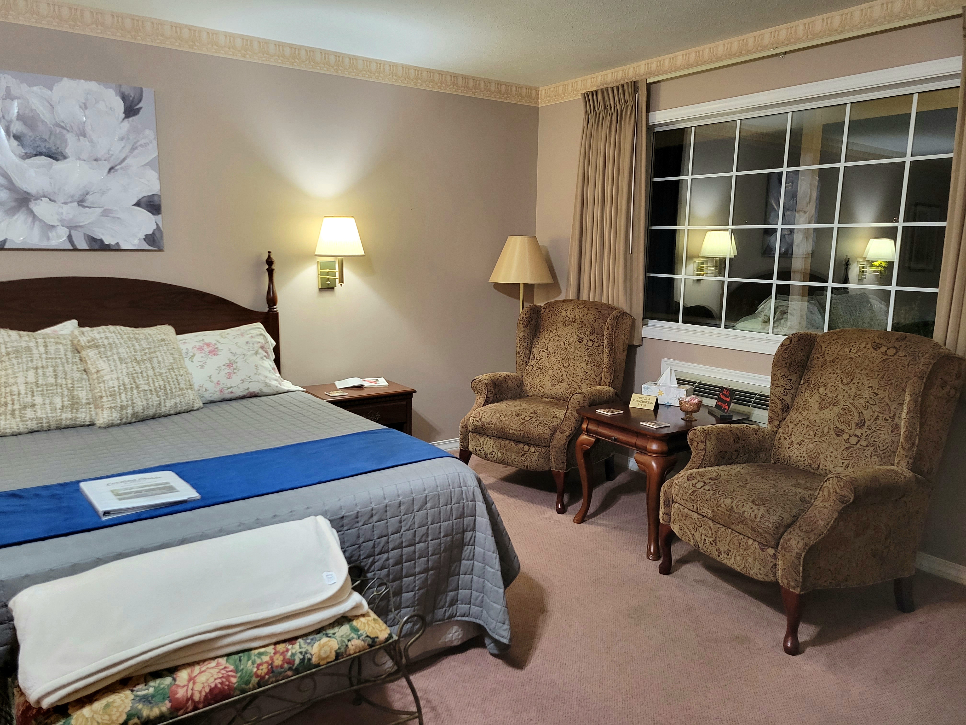 An interior view of a cozy seating area featuring two upholstered wingback chairs positioned next to a large window. A small wooden side table sits between the chairs, and a king-size bed with a blue runner is visible to the left.