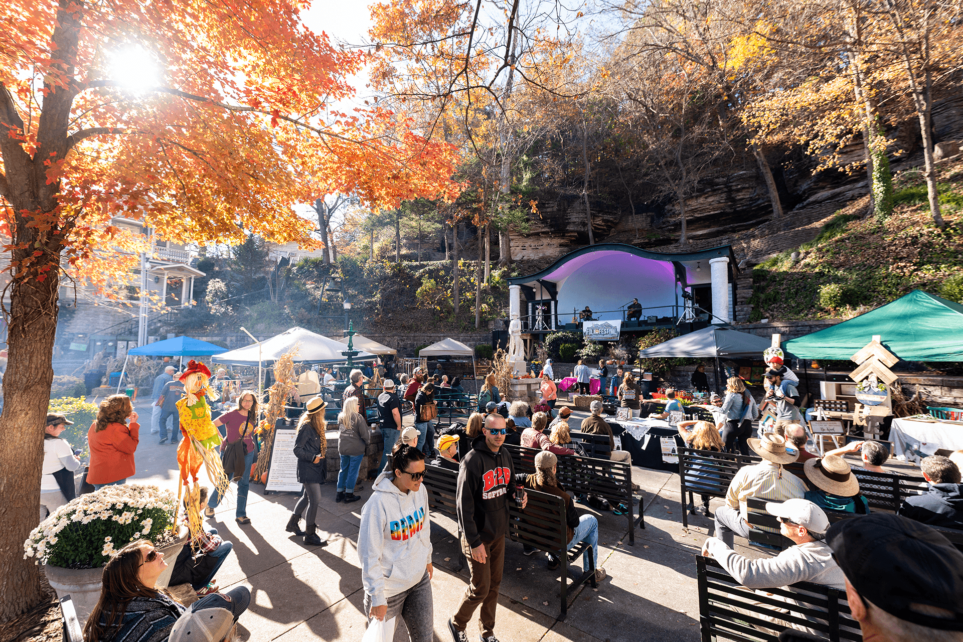 A vibrant outdoor festival scene with people gathered around a stage, surrounded by colorful fall foliage and vendor tents.