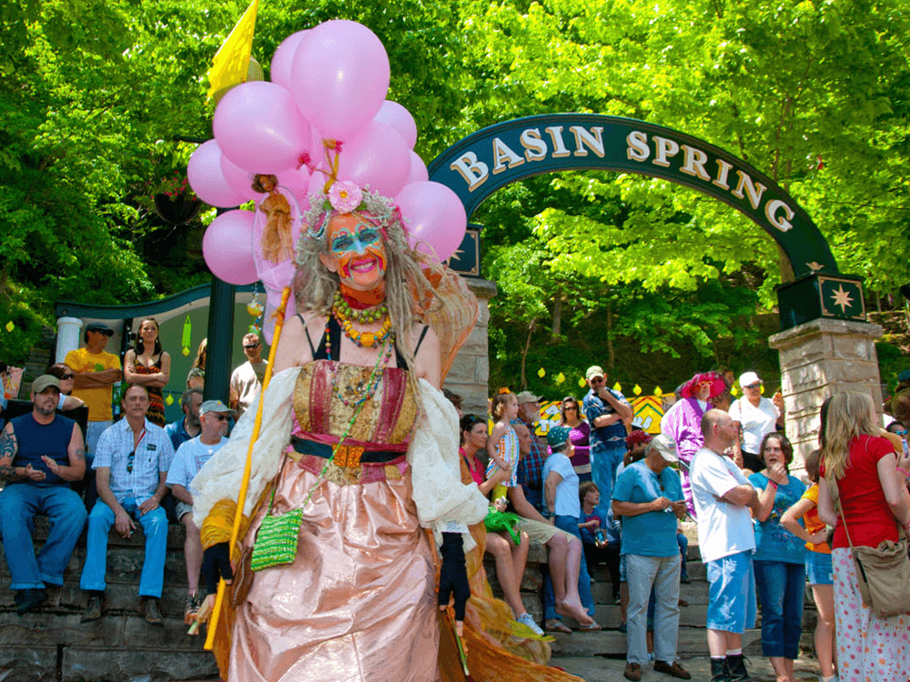 A colorful figure in costume holding pink balloons walks in front of a crowd at Basin Spring.