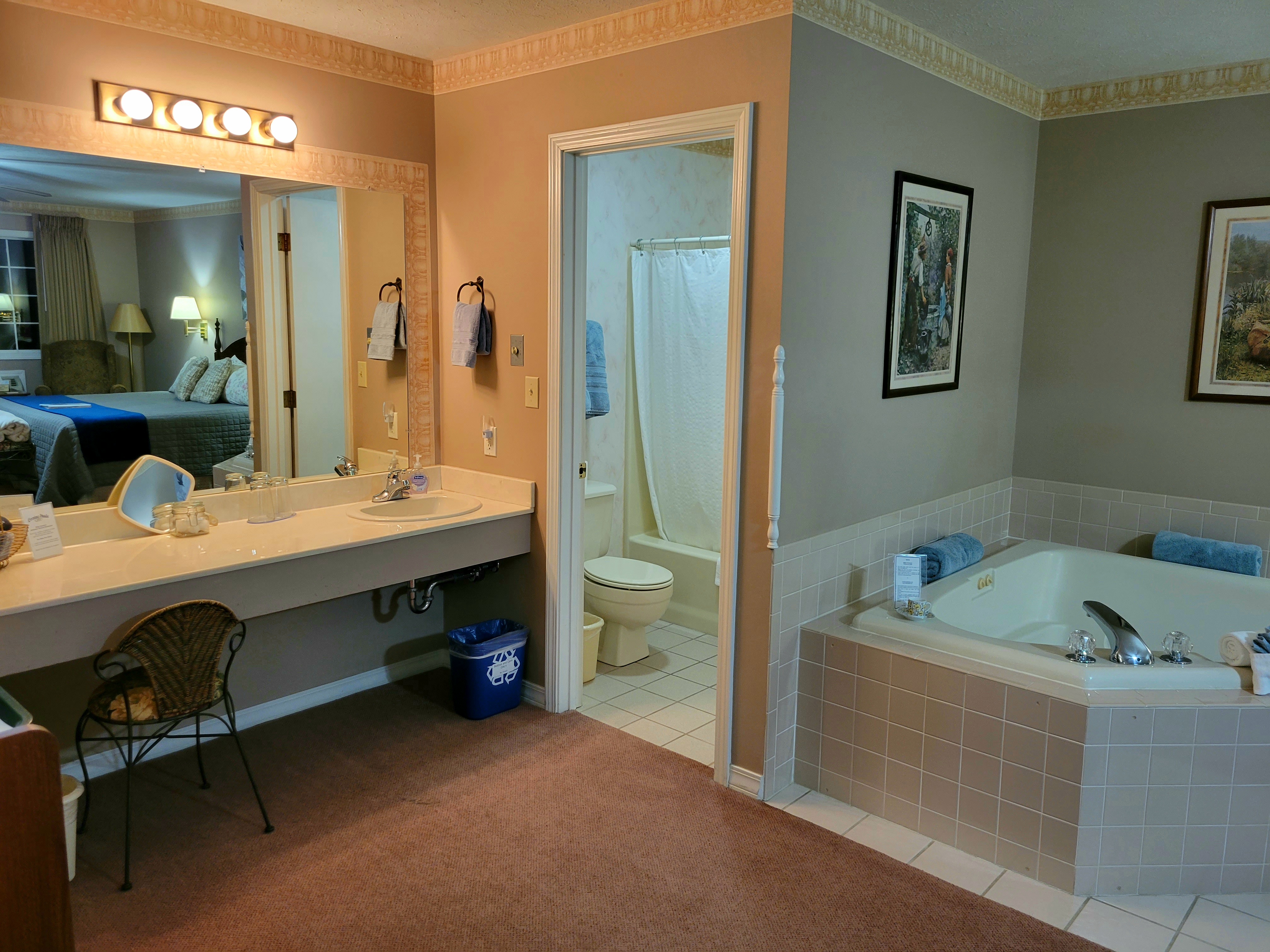 A spacious suite showing a long vanity with a large mirror that reflects the king-size bed. To the right, a large jetted tub is built into the corner, and a doorway leads into a separate bathroom area with a toilet and shower.