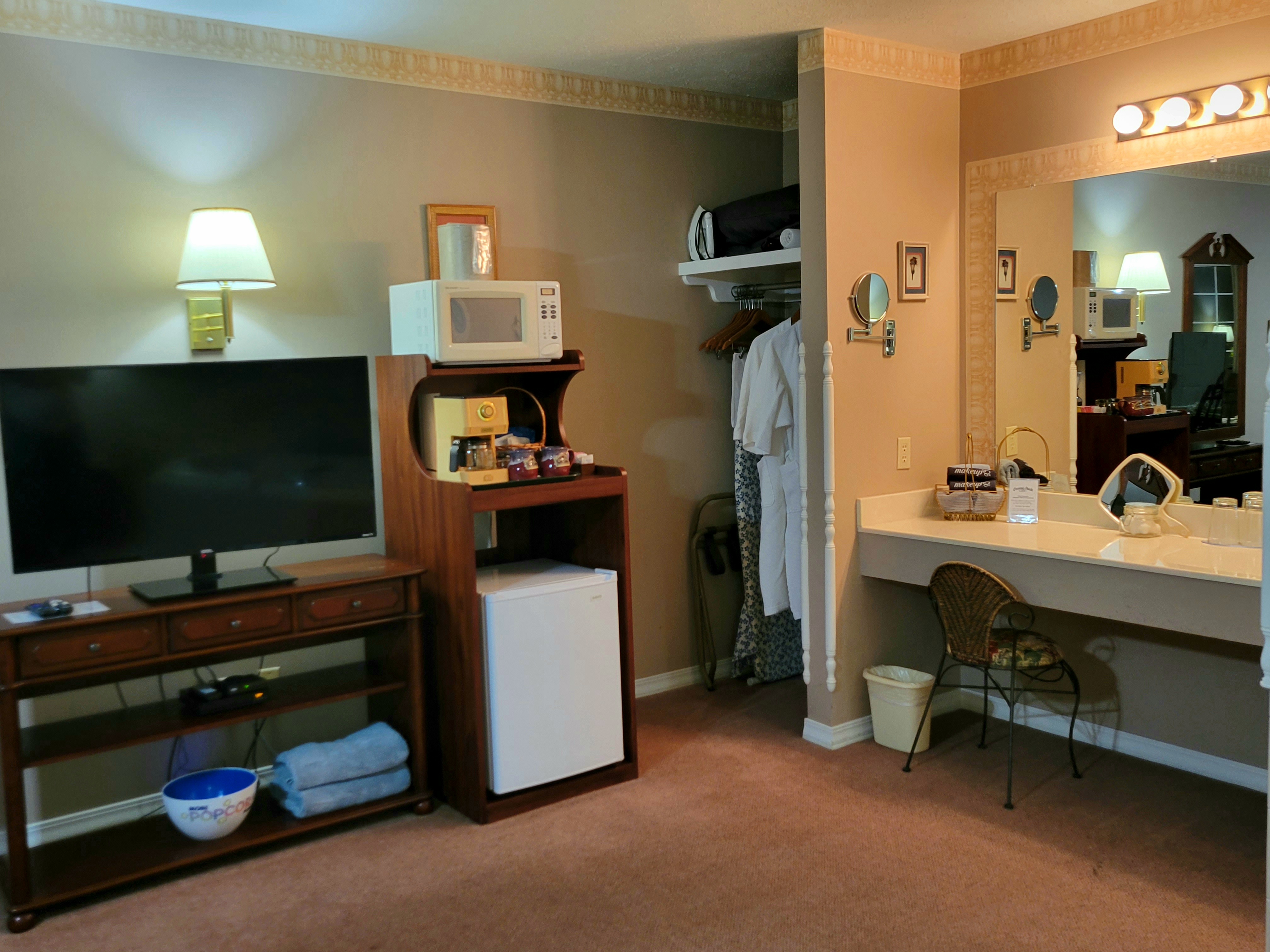 An interior view of a suite showing a entertainment and vanity area. It features a flat-screen TV on a wooden stand, a refreshment station with a microwave, coffee maker, and mini-fridge, and a large lit vanity mirror with a desk chair.