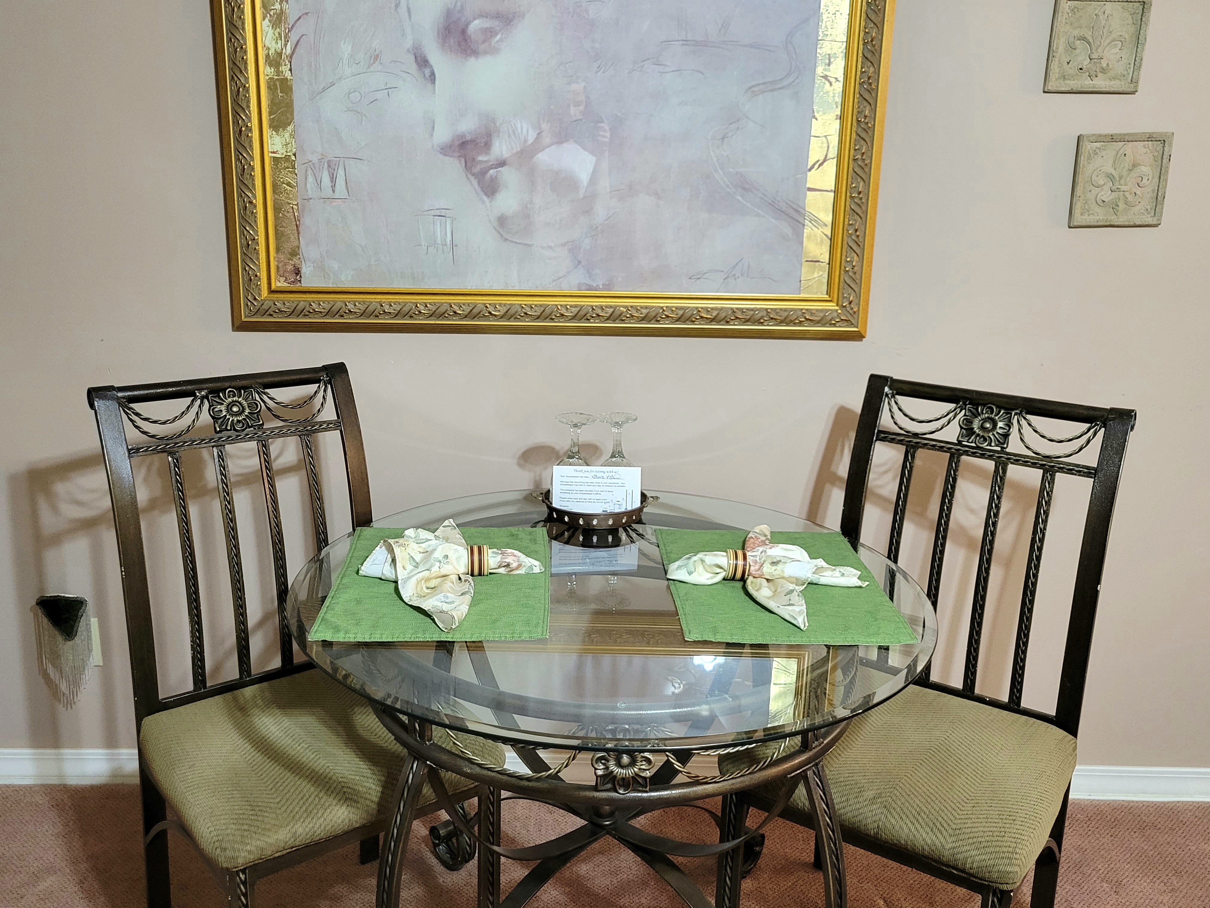 A glass-top dining table with two metal-frame chairs, set with green placemats and patterned napkins. A large gold-framed piece of artwork hangs on the wall behind the table.