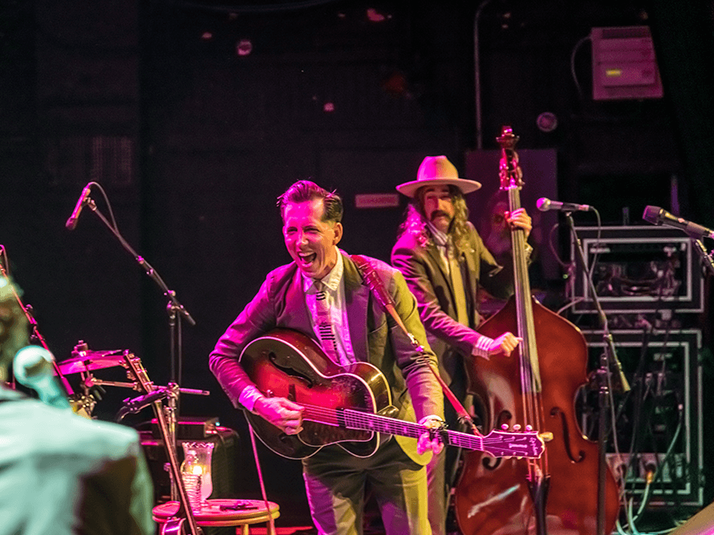 A musician joyfully plays an acoustic guitar on stage while a bassist stands in the background.