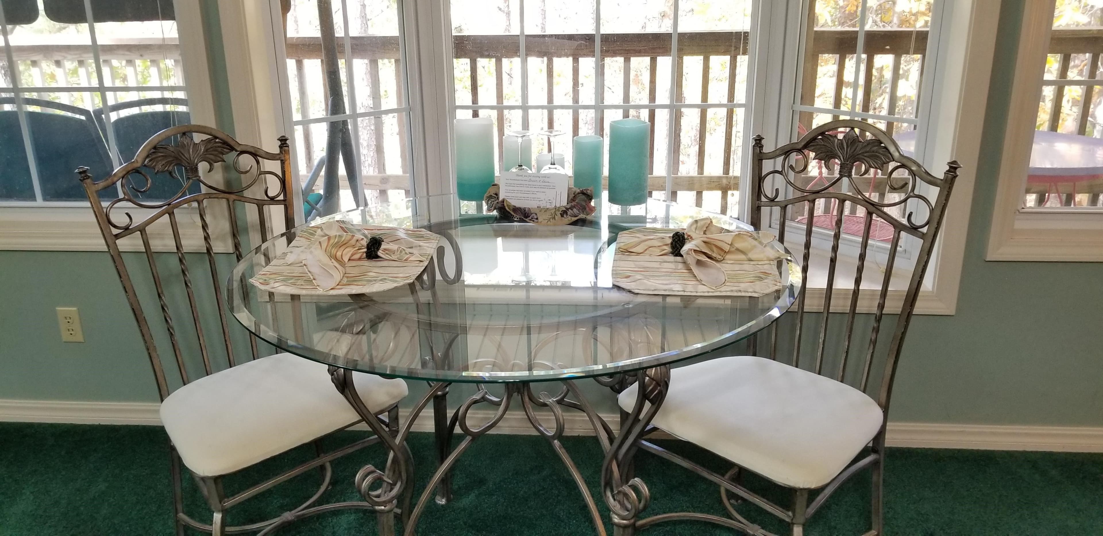 2 Person dining table with a glass table top and iron chairs with white cushioned seats. There are faux candles on the table, wine glasses, and napkins atop of matching floral placemats. There is a bay window with a window seat behind the table. The walls inside the room are light blue and the carpet is dark green. Through the open windows one can see the deck area out front.