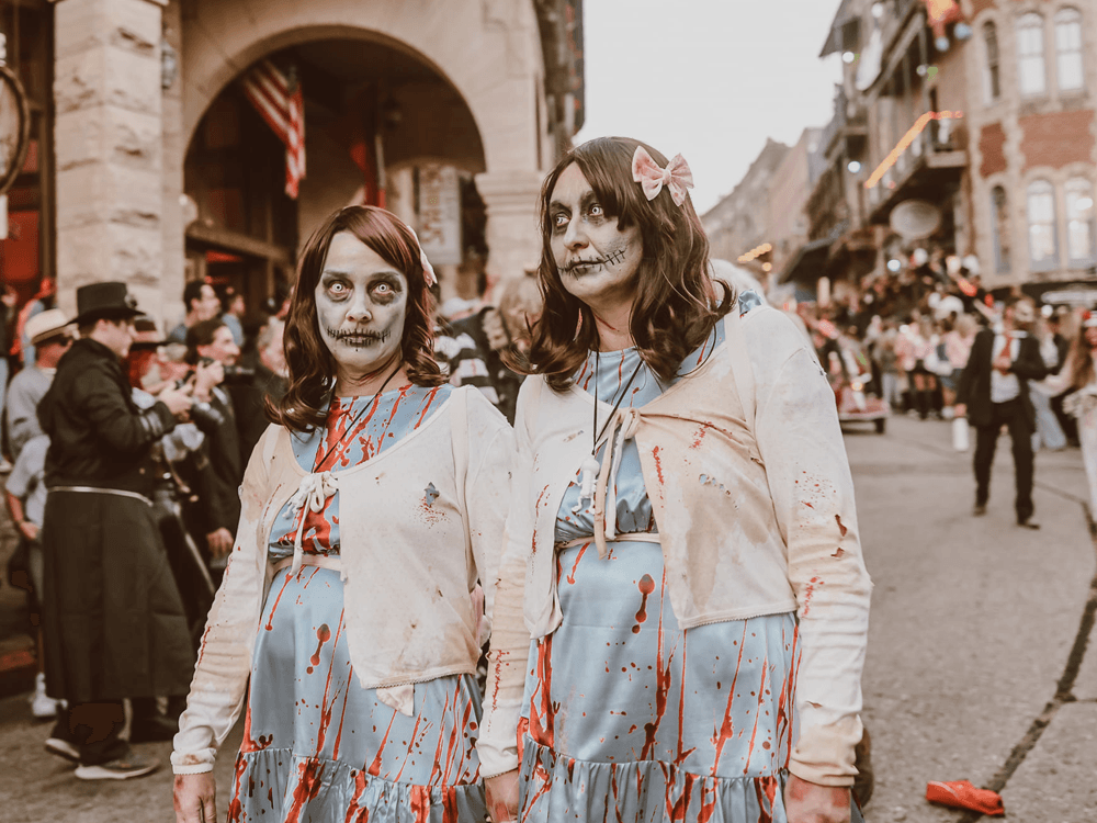 Two zombie women in tattered, blood-stained dresses stand on a festive street during a parade.