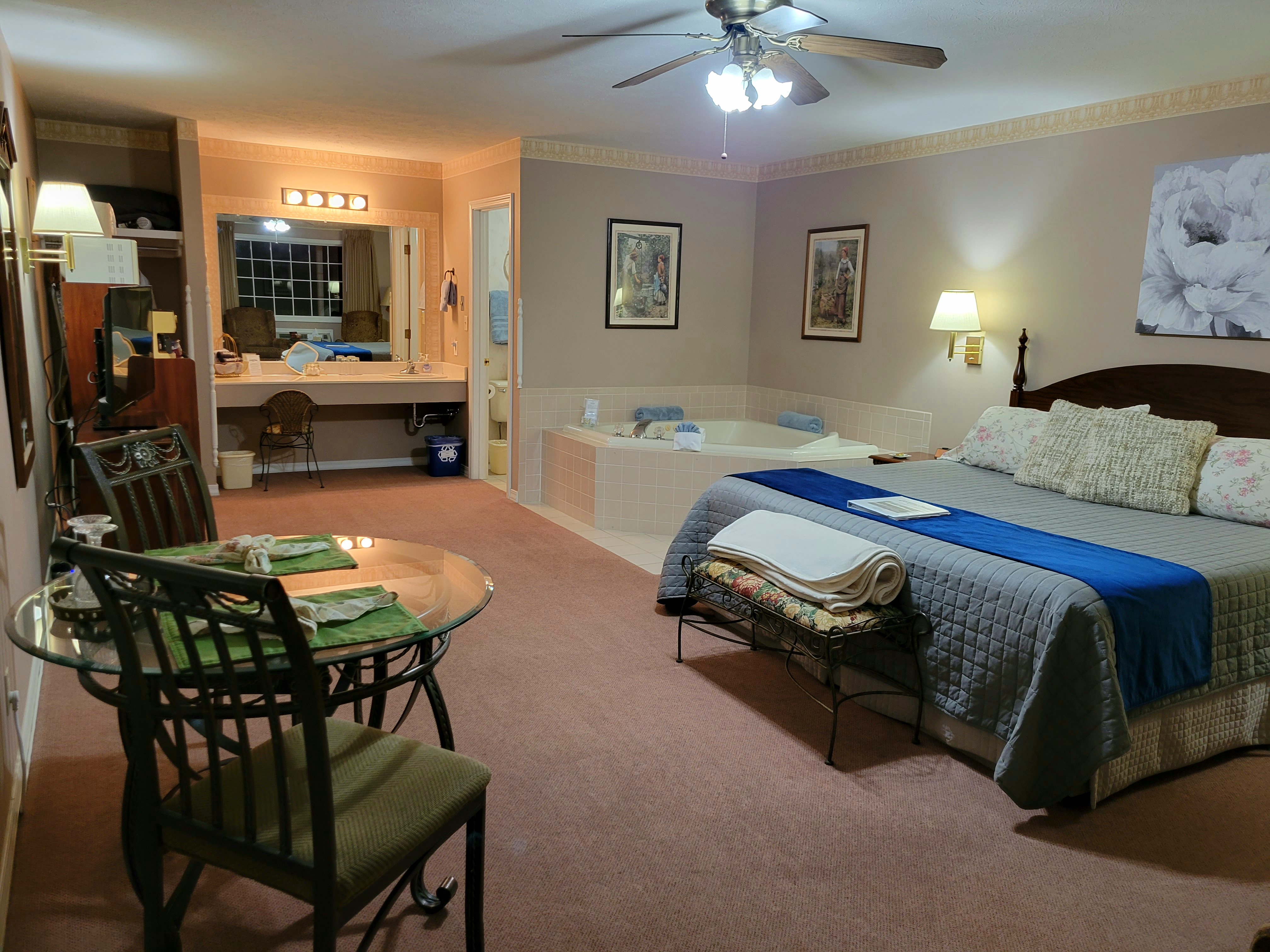 A wide-angle interior view of a suite featuring a glass-top dining table with two chairs in the foreground. The room includes a king-size bed with a blue runner, a large corner jetted tub, and a vanity area with a large mirror in the background.