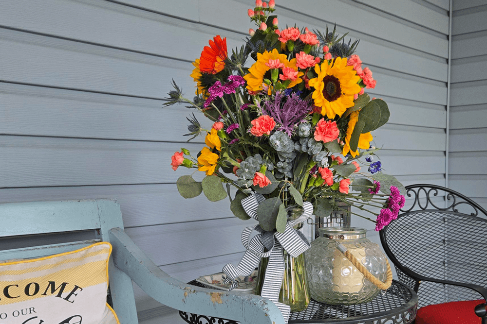 A vibrant bouquet of flowers, including sunflowers and pink carnations, sits on a table next to a cozy chair on a porch.