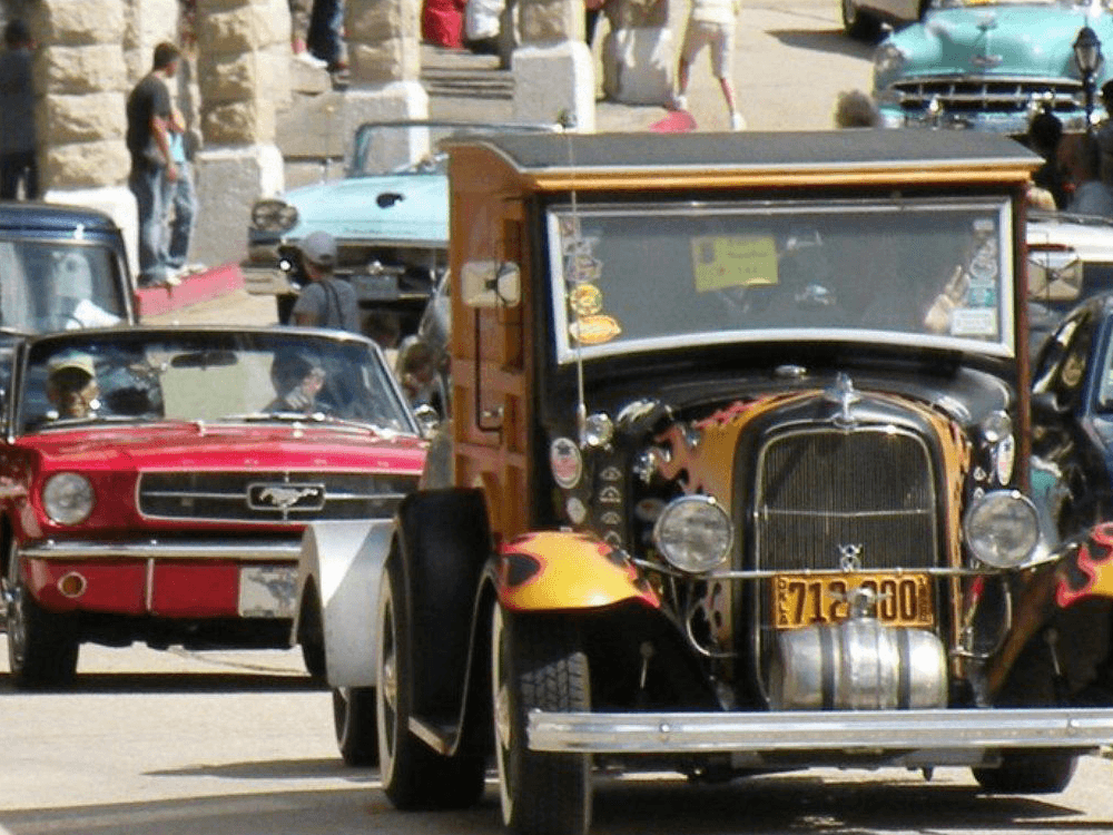 A vintage orange hot rod with flames and a classic red convertible are seen on a bustling street filled with cars and people.