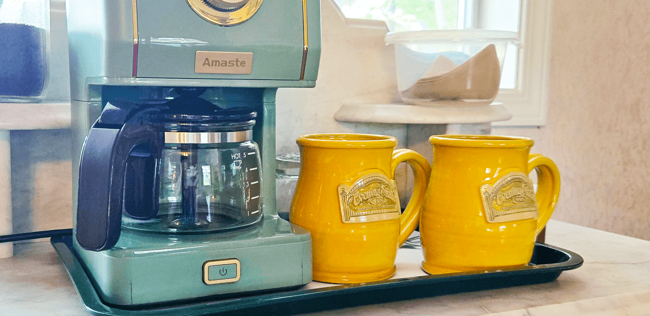 A retro-style teal coffee maker with a glass pot sits on a countertop next to two bright yellow mugs. The scene feels cozy and inviting.