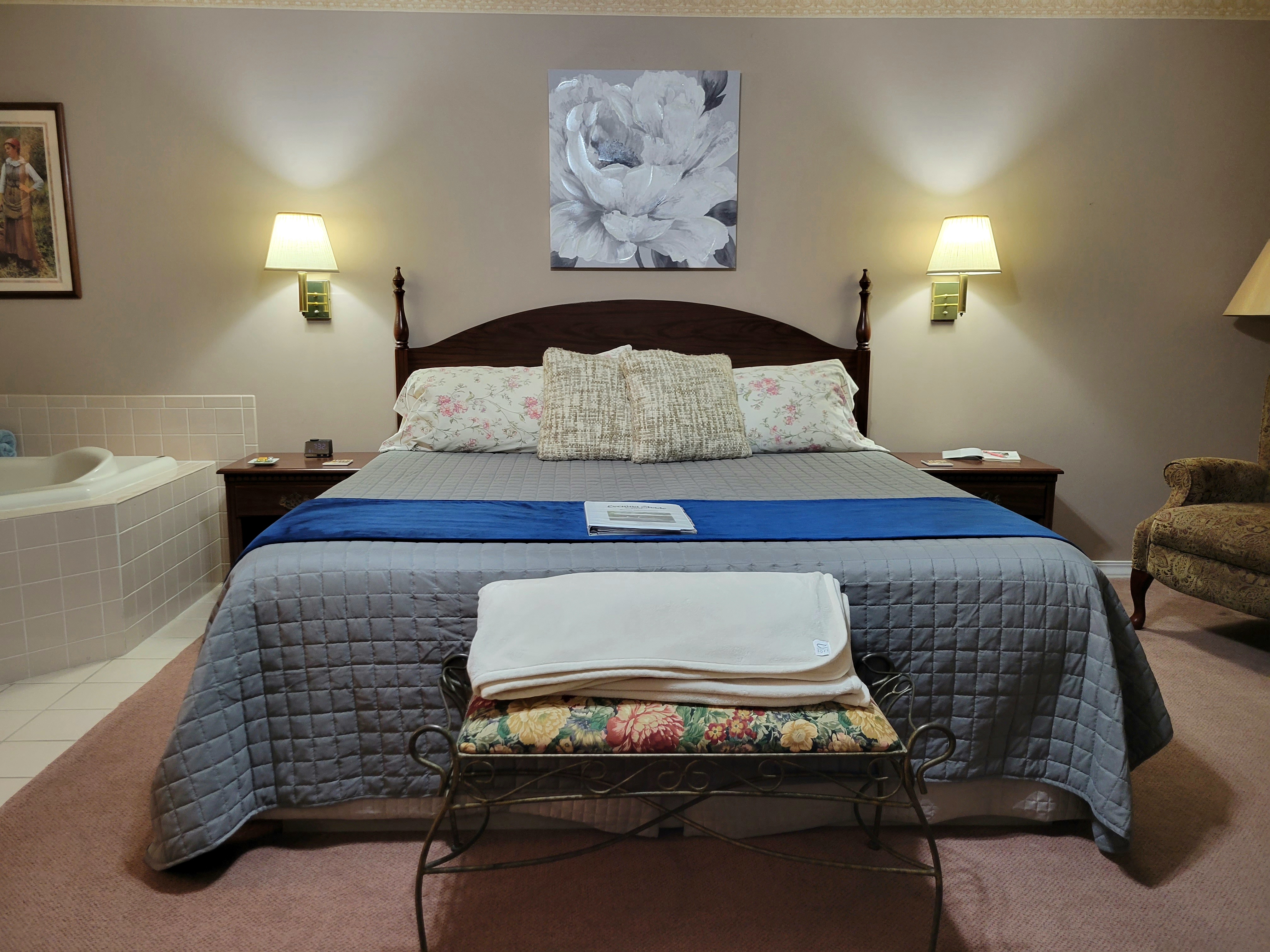 An interior view of a bedroom featuring a king-size bed with a blue runner, framed by two wooden nightstands with wall-mounted lamps. A large jetted tub is built into the corner to the left, and a floral painting hangs above the headboard.