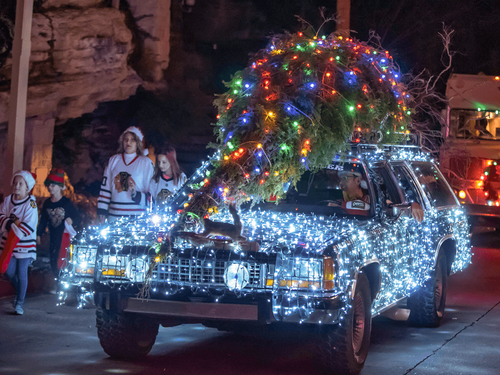 A festive car adorned with colorful lights and a large decorated tree is featured in a nighttime holiday parade.