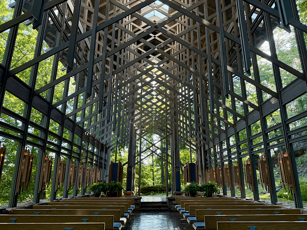 A modern chapel with a geometric wooden structure and rows of benches, surrounded by greenery.