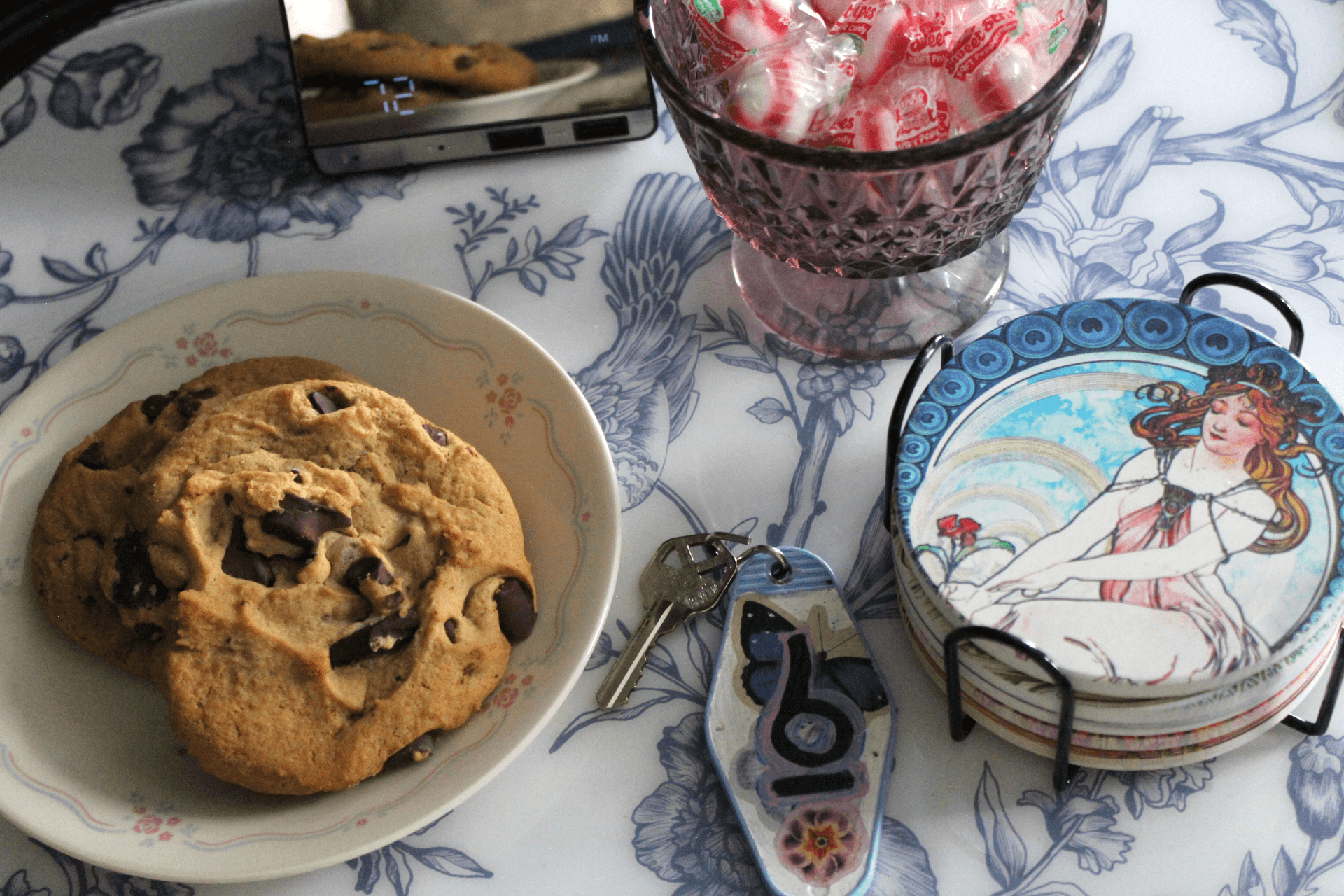 A plate of chocolate chip cookies, a glass bowl of mints, a set of illustrated coasters, house keys, and a phone rest on a floral-patterned surface.