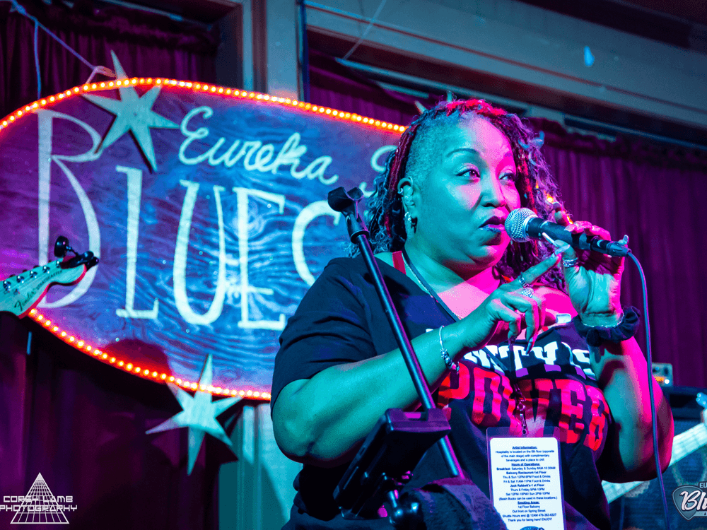 A woman speaks into a microphone on stage in front of a brightly lit "Eureka Springs Blues" sign.