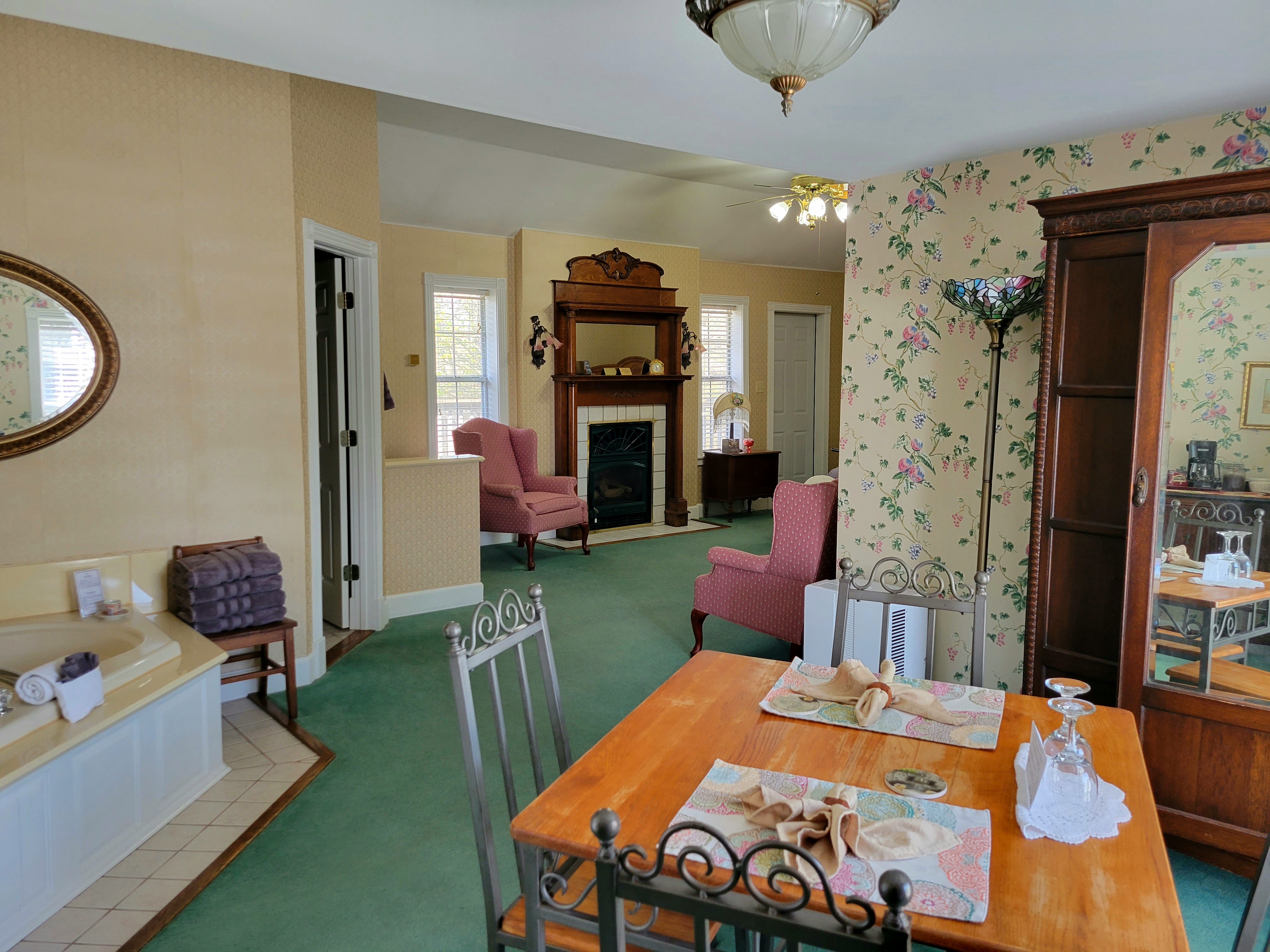 A spacious suite featuring a wooden dining table in the foreground. The open-plan room extends to a cozy seating area with two pink wingback chairs, a classic fireplace with a detailed wooden mantel, and a corner jetted tub partially visible to the left.