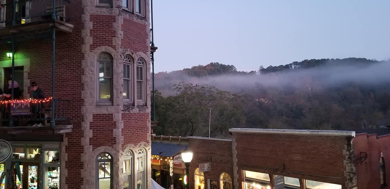 Red brick building with arched windows, glowing shop lights, and misty hills in the background. Early evening ambiance with a cozy, quaint feel.