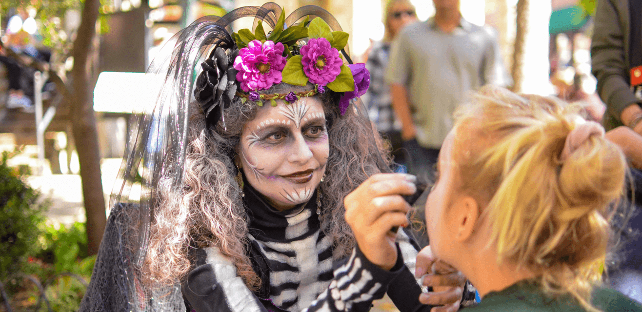 A performer in elaborate skeleton makeup applies face paint to a woman during a festive event.